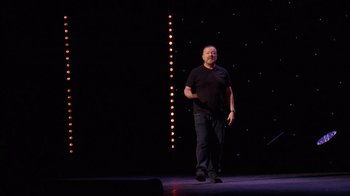 Movie still from “Ricky Gervais: SuperNature” (2022), directed by John L. Spencer – A man standing on a stage in front of lights; Wide shot, Low angle
