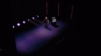 Movie still from “Ricky Gervais: SuperNature” (2022), directed by John L. Spencer – A man standing on a stage in front of a microphone; Extreme Wide shot, Overhead angle