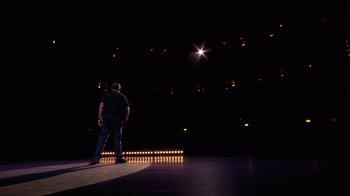 Movie still from “Ricky Gervais: SuperNature” (2022), directed by John L. Spencer – A man standing on a stage in the middle of a dark room; Extreme Wide shot, High angle