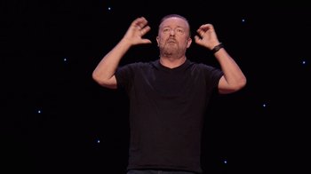 Movie still from “Ricky Gervais: SuperNature” (2022), directed by John L. Spencer – A man in a black t - shirt is holding his hands up in the air; Medium shot, Overhead angle
