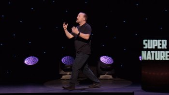Movie still from “Ricky Gervais: SuperNature” (2022), directed by John L. Spencer – A man standing on a stage in front of a microphone; Wide shot, Low angle