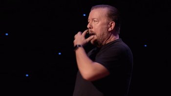 Movie still from “Ricky Gervais: SuperNature” (2022), directed by John L. Spencer – A man with a beard and a black t - shirt is on a stage; Close Up shot, Over the shoulder angle