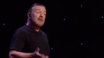 Movie still from “Ricky Gervais: SuperNature” (2022), directed by John L. Spencer – A man with a goatee is talking on stage; Close Up shot, Overhead angle