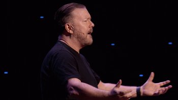 Movie still from “Ricky Gervais: SuperNature” (2022), directed by John L. Spencer – A man with a beard is talking on a stage; Close Up shot, High angle