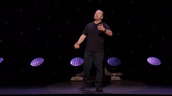 Movie still from “Ricky Gervais: SuperNature” (2022), directed by John L. Spencer – A man standing on a stage in front of a microphone; Medium shot, Over the shoulder angle