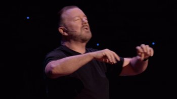 Movie still from “Ricky Gervais: SuperNature” (2022), directed by John L. Spencer – A man in a black t - shirt is holding his hands up; Close Up shot, Overhead angle