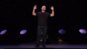 Movie still from “Ricky Gervais: SuperNature” (2022), directed by John L. Spencer – A man standing on a stage with his hands raised; Wide shot, Low angle