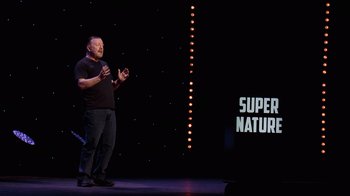 Movie still from “Ricky Gervais: SuperNature” (2022), directed by John L. Spencer – A man standing on a stage in front of a microphone; Medium shot, Over the shoulder angle