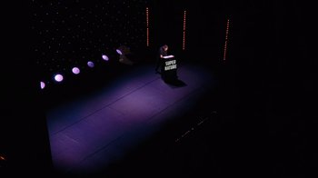 Movie still from “Ricky Gervais: SuperNature” (2022), directed by John L. Spencer – A man standing on a stage holding a microphone; Extreme Wide shot, High angle