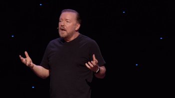 Movie still from “Ricky Gervais: SuperNature” (2022), directed by John L. Spencer – An image of a man giving a speech on a stage; Medium shot, Overhead angle