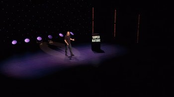 Movie still from “Ricky Gervais: SuperNature” (2022), directed by John L. Spencer – A man standing on a stage in front of a microphone; Extreme Wide shot, High angle