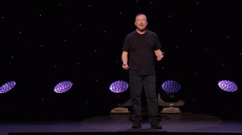 Movie still from “Ricky Gervais: SuperNature” (2022), directed by John L. Spencer – A man standing on a stage in front of purple lights; Medium shot, Over the shoulder angle