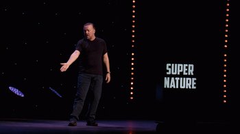 Movie still from “Ricky Gervais: SuperNature” (2022), directed by John L. Spencer – A man standing on a stage holding his hand out to someone; Wide shot, Over the shoulder angle