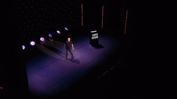 Movie still from “Ricky Gervais: SuperNature” (2022), directed by John L. Spencer – A man standing on a stage in front of a microphone; Extreme Wide shot, High angle