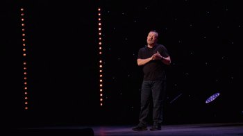 Movie still from “Ricky Gervais: SuperNature” (2022), directed by John L. Spencer – A man standing on a stage in front of a microphone; Medium shot, Low angle
