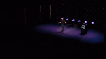 Movie still from “Ricky Gervais: SuperNature” (2022), directed by John L. Spencer – A man standing on a stage in front of a microphone; Extreme Wide shot, High angle