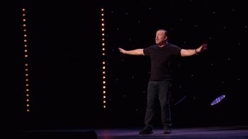 Movie still from “Ricky Gervais: SuperNature” (2022), directed by John L. Spencer – A man standing on a stage with his arms outstretched; Wide shot, Low angle
