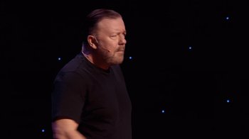 Movie still from “Ricky Gervais: SuperNature” (2022), directed by John L. Spencer – A man in a black shirt is standing in front of a black background; Close Up shot, Over the shoulder angle