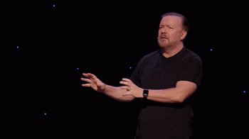 Movie still from “Ricky Gervais: SuperNature” (2022), directed by John L. Spencer – A man standing on a stage in front of a microphone; Medium shot, High angle