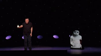 Movie still from “Ricky Gervais: SuperNature” (2022), directed by John L. Spencer – A man standing on a stage in front of an image of a baby; Medium shot, High angle