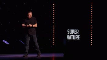 Movie still from “Ricky Gervais: SuperNature” (2022), directed by John L. Spencer – A man standing on a stage holding a microphone; Wide shot, Low angle