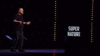 Movie still from “Ricky Gervais: SuperNature” (2022), directed by John L. Spencer – A man standing on a stage holding a blue object; Wide shot, Over the shoulder angle