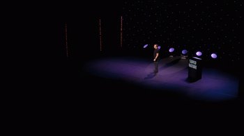 Movie still from “Ricky Gervais: SuperNature” (2022), directed by John L. Spencer – A man standing on a stage holding a frisbee in his hand; Extreme Wide shot, High angle