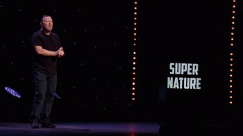 Movie still from “Ricky Gervais: SuperNature” (2022), directed by John L. Spencer – A man standing on a stage holding a microphone; Extreme Wide shot, Over the shoulder angle