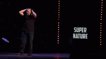 Movie still from “Ricky Gervais: SuperNature” (2022), directed by John L. Spencer – A man standing on a stage holding his hands behind his head; Medium shot, Over the shoulder angle