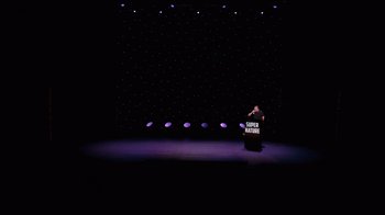 Movie still from “Ricky Gervais: SuperNature” (2022), directed by John L. Spencer – A man standing at a podium on a stage; Extreme Wide shot, High angle