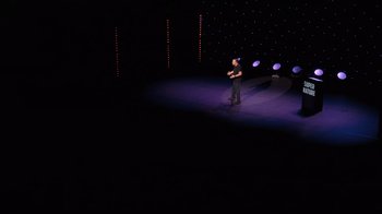 Movie still from “Ricky Gervais: SuperNature” (2022), directed by John L. Spencer – A man standing on a stage in front of a microphone; Extreme Wide shot, High angle