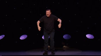 Movie still from “Ricky Gervais: SuperNature” (2022), directed by John L. Spencer – A man standing on a stage in front of a black background; Wide shot, High angle