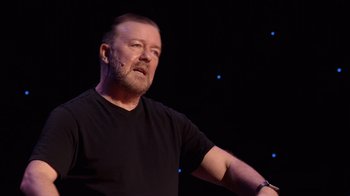 Movie still from “Ricky Gervais: SuperNature” (2022), directed by John L. Spencer – A man in a black t - shirt is holding his arm up; Close Up shot, High angle