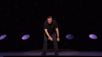 Movie still from “Ricky Gervais: SuperNature” (2022), directed by John L. Spencer – A man standing on a stage in front of a purple background; Wide shot, High angle