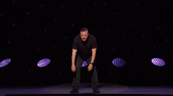 Movie still from “Ricky Gervais: SuperNature” (2022), directed by John L. Spencer – A man standing on a stage in front of purple lights; Medium shot, High angle