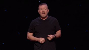 Movie still from “Ricky Gervais: SuperNature” (2022), directed by John L. Spencer – A man in a black t - shirt is giving a speech; Medium shot, High angle
