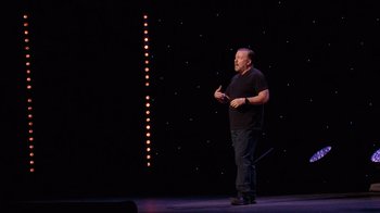 Movie still from “Ricky Gervais: SuperNature” (2022), directed by John L. Spencer – A man standing on a stage in front of a microphone; Medium shot, Low angle