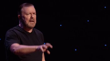 Movie still from “Ricky Gervais: SuperNature” (2022), directed by John L. Spencer – A man in a black t - shirt talking on a stage; Close Up shot, High angle