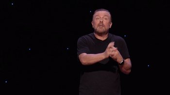 Movie still from “Ricky Gervais: SuperNature” (2022), directed by John L. Spencer – A man in a black t - shirt is clapping his hands; Medium shot, Overhead angle
