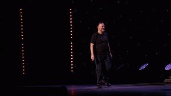 Movie still from “Ricky Gervais: SuperNature” (2022), directed by John L. Spencer – A man standing on a stage holding a wand; Wide shot, Low angle