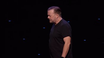 Movie still from “Ricky Gervais: SuperNature” (2022), directed by John L. Spencer – A man in a black t - shirt is looking down; Medium shot, Low angle