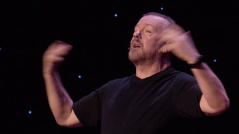 Movie still from “Ricky Gervais: SuperNature” (2022), directed by John L. Spencer – A man in a black t - shirt is holding his hands in the air; Close Up shot, Low angle