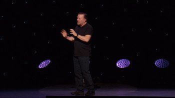 Movie still from “Ricky Gervais: SuperNature” (2022), directed by John L. Spencer – A man standing on a stage in front of a microphone; Medium shot, Low angle