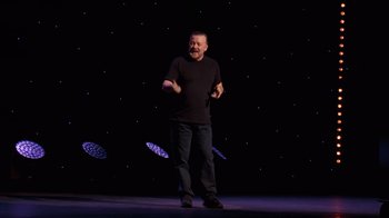 Movie still from “Ricky Gervais: SuperNature” (2022), directed by John L. Spencer – A man standing on a stage in front of a black background; Wide shot, Low angle