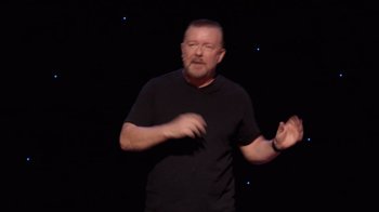 Movie still from “Ricky Gervais: SuperNature” (2022), directed by John L. Spencer – A man in a black t - shirt is giving a speech; Medium shot, High angle