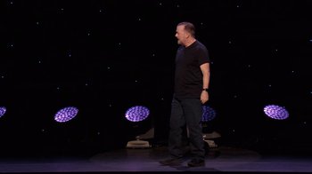 Movie still from “Ricky Gervais: SuperNature” (2022), directed by John L. Spencer – A man standing on a stage in front of a microphone; Wide shot, Over the shoulder angle