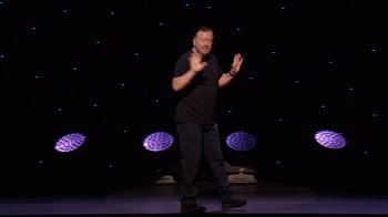 Movie still from “Ricky Gervais: SuperNature” (2022), directed by John L. Spencer – A man standing on a stage in front of a microphone; Wide shot, High angle