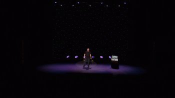 Movie still from “Ricky Gervais: SuperNature” (2022), directed by John L. Spencer – A man standing in front of a microphone on a stage; Extreme Wide shot, Low angle