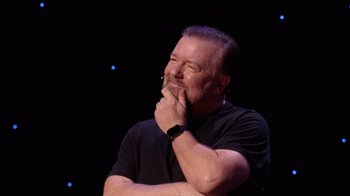 Movie still from “Ricky Gervais: SuperNature” (2022), directed by John L. Spencer – A man with a beard and a black t - shirt is holding his hand to his face; Close Up shot, Over the shoulder angle