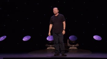Movie still from “Ricky Gervais: SuperNature” (2022), directed by John L. Spencer – A man standing on a stage in front of lights; Wide shot, Low angle
